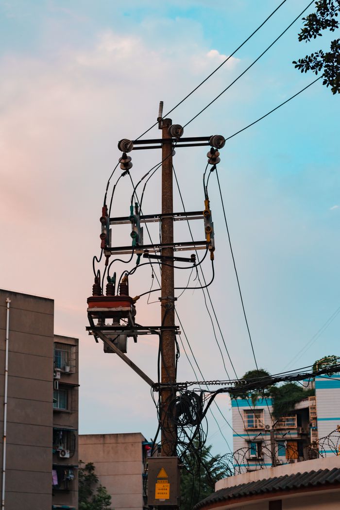 A tall utility pole with complex electrical wiring set against a colorful evening sky in an urban area.